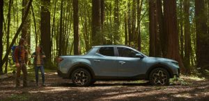 A light blue 2022 Hyundai Santa Cruz parked in a heavily wooded area with a couple standing behind it. | Hyundai dealer in Little Rock, AR.